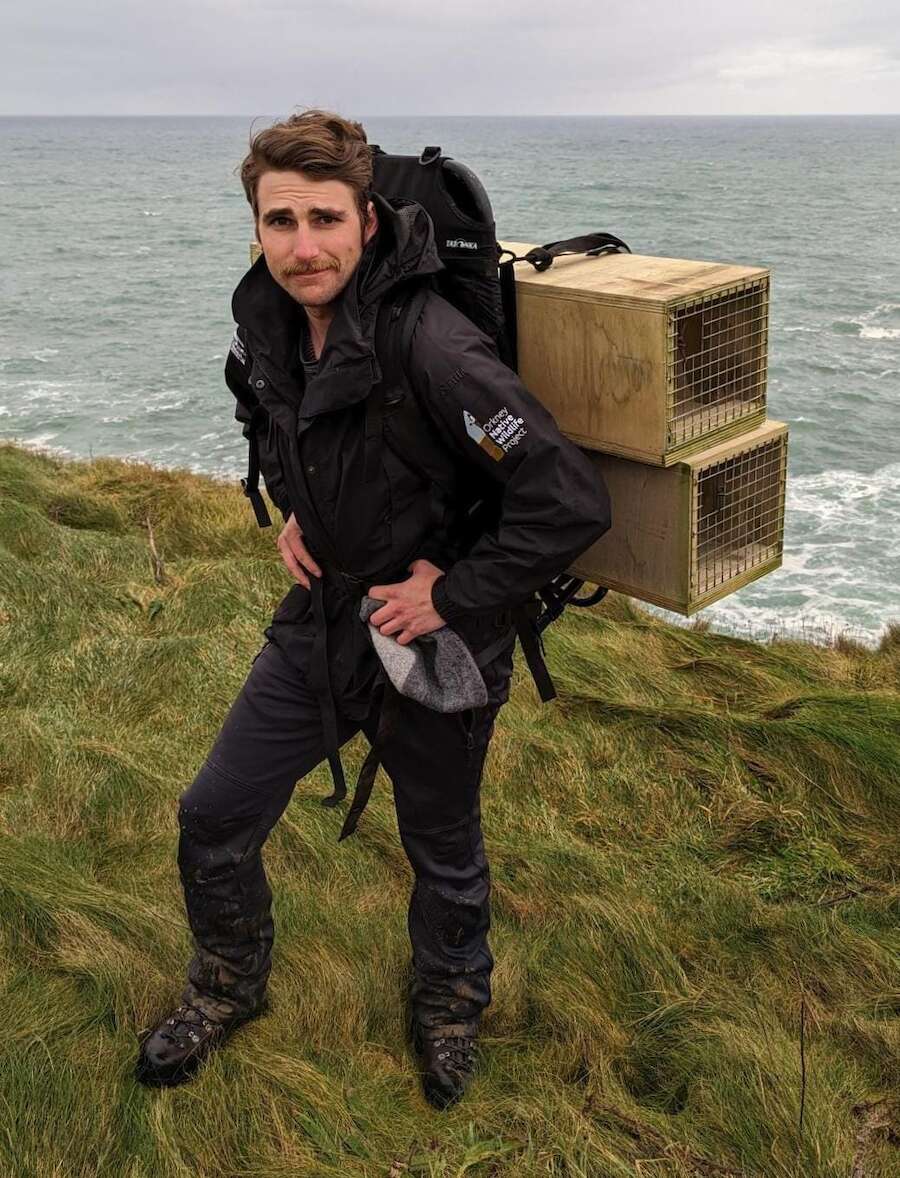 Denis stands on a clifftop kitted out all in black with two wooden trap boxes on his back, a wooly hat in hand and a smirk on his face