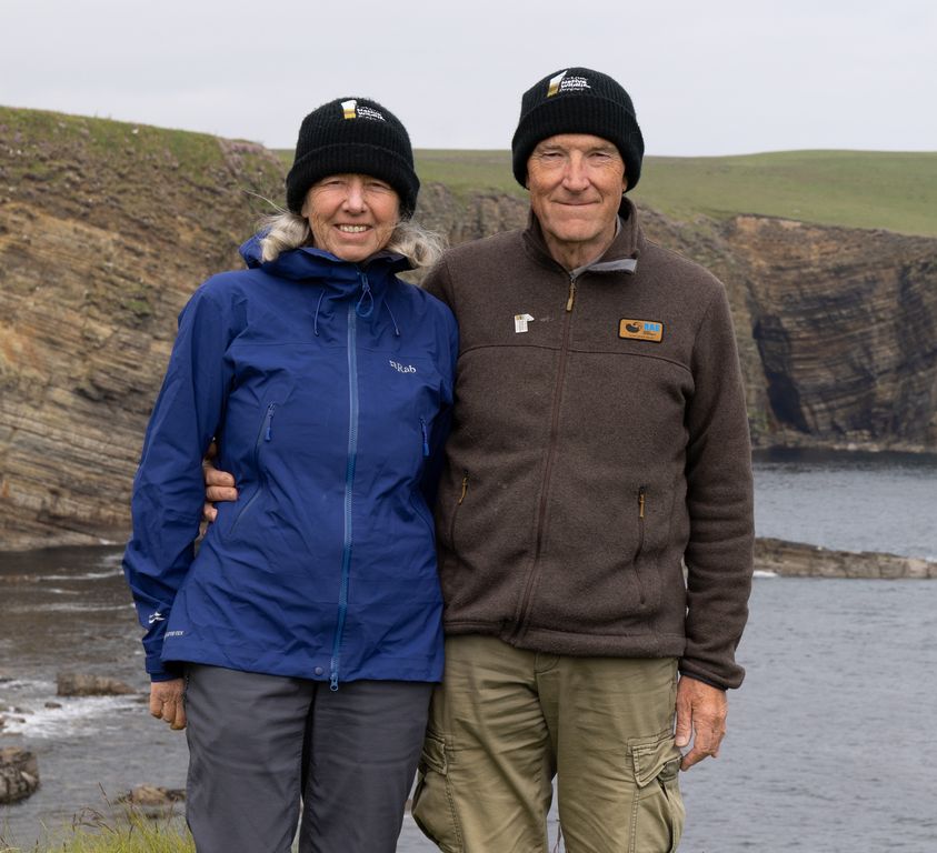 Sandie and Mike stand side by side facing the camera with a cliff and the sea behind. Mike has his arm around Sandie's waist. Both wear black hats with the project logo on and outdoor clothing.