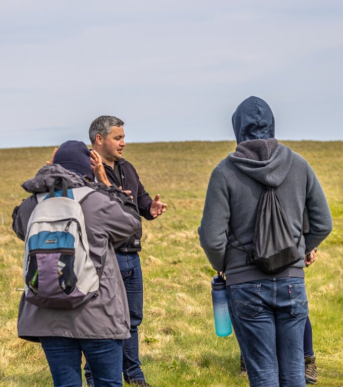 A project staff member stands in the background facing the camera talking to two people with their backs to the camera